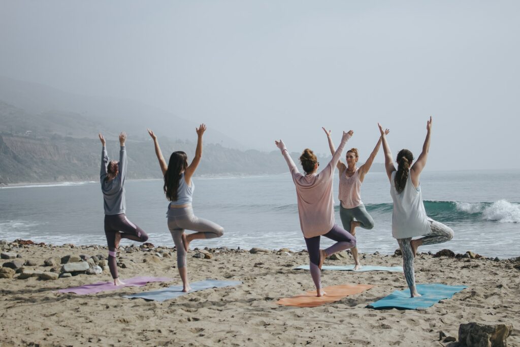 wellness trends - corporate yoga - team of woman in tree pose standing on the shore line of a beach