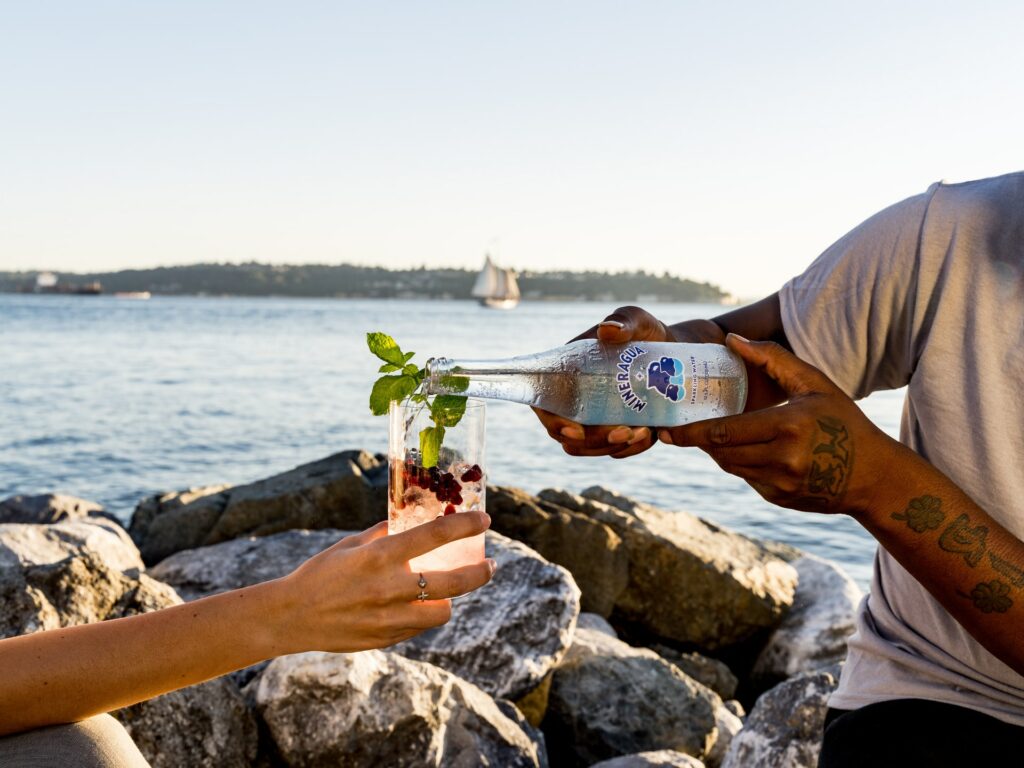 wellness trend 2022 - sober curious - two people sitting by the water, one pouring sparkling water into a glass held by another person.